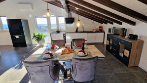 A modern kitchen with a dining area, featuring a large island with a meal set, pendant lighting, and a view of the outdoors. - Cool 3-bedroom house with AC in charming Praputnjak (Hreljin)