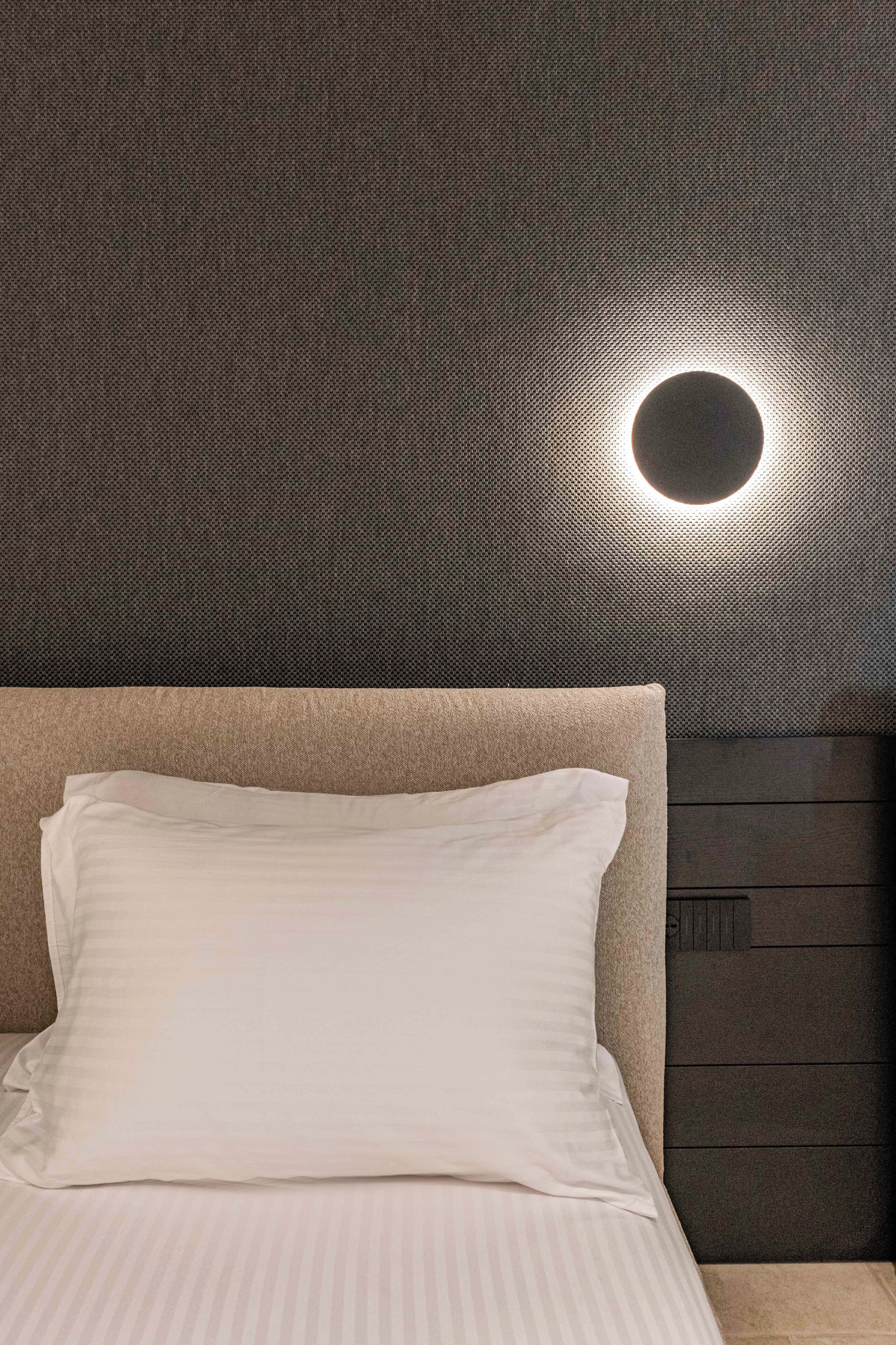 Un lit bien fait, doté d’une tête de lit, d’un luminaire circulaire fixé au mur et d’un panneau en bois foncé sur le mur.
