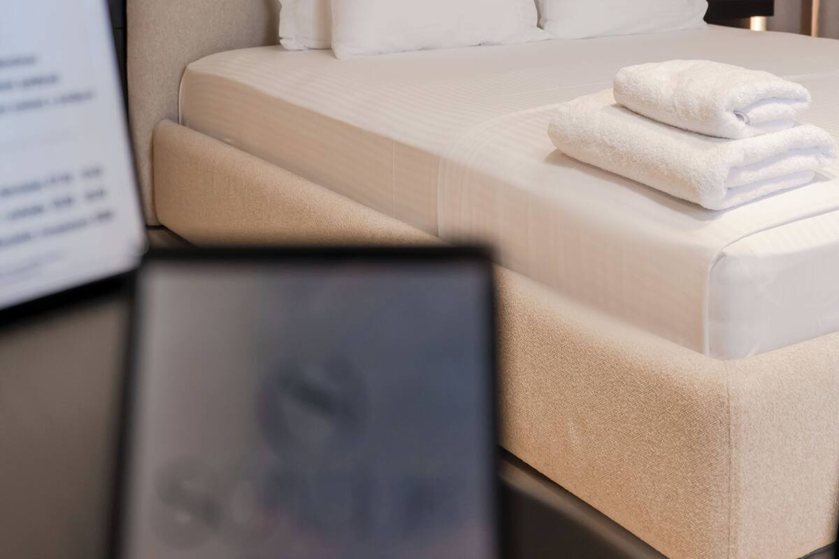 A neatly made bed with white linens and pillows, a bedside table with a lamp, and a framed picture on the wall.