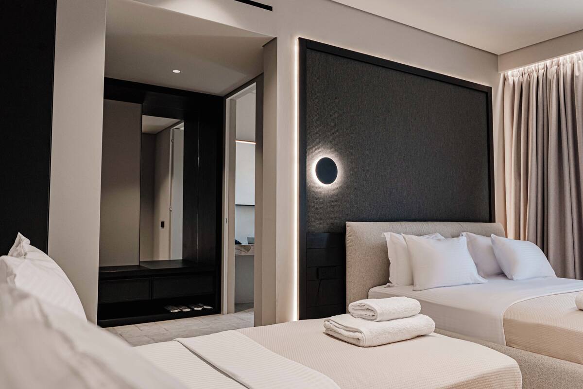 A modern hotel room with a large bed, a smaller bed, and a seating area. There is a bathroom visible in the background.