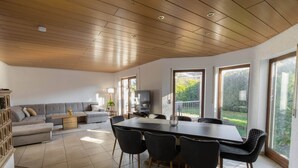 Un vaste espace de vie doté d’un plafond en bois, d’une grande table à manger avec des chaises, d’un canapé et d’un téléviseur.