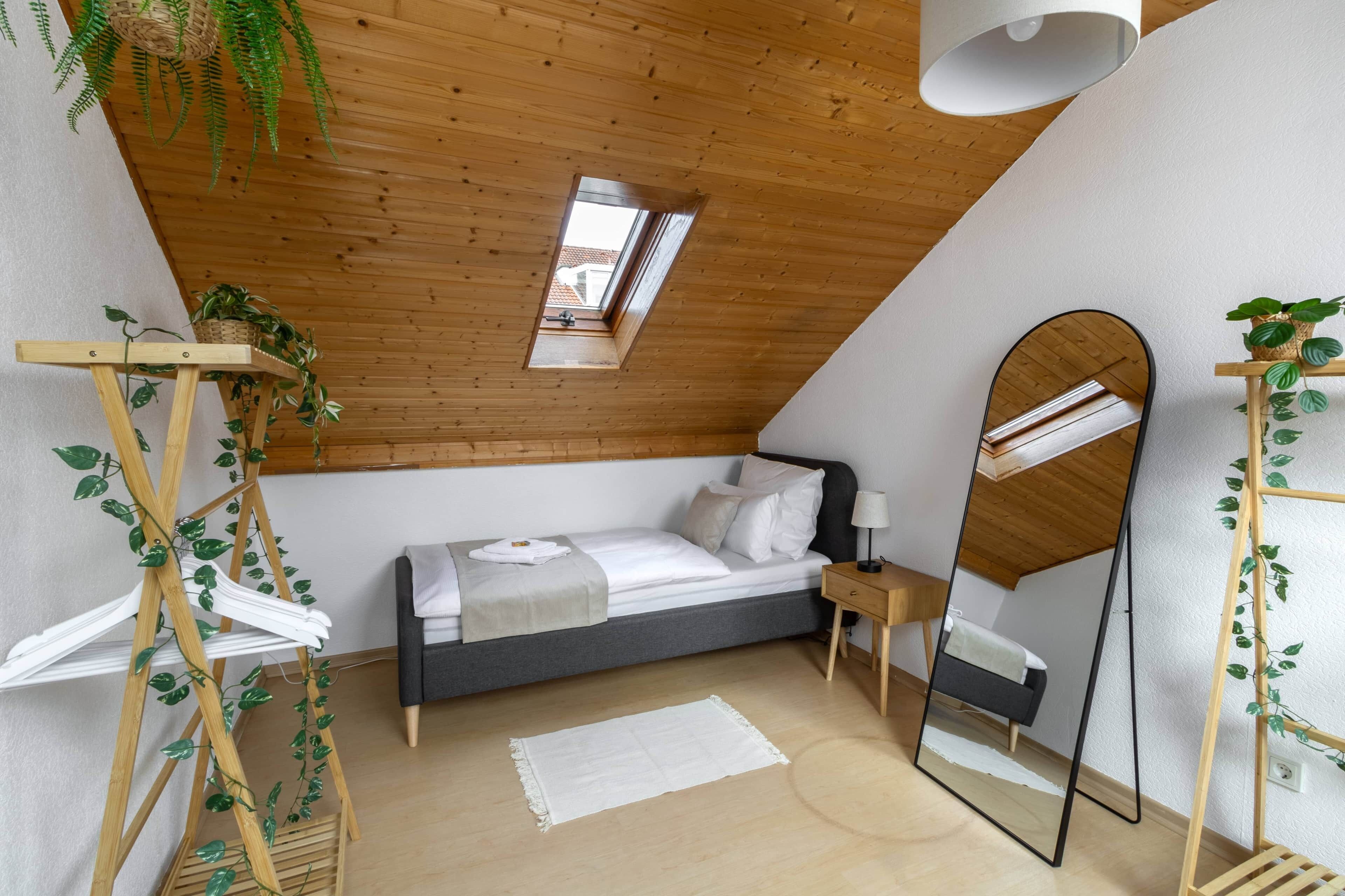 Une chambre avec un plafond en pente, un puits de lumière, un lit avec des oreillers, une table de chevet avec une lampe, un miroir et un support à plantes en bois.