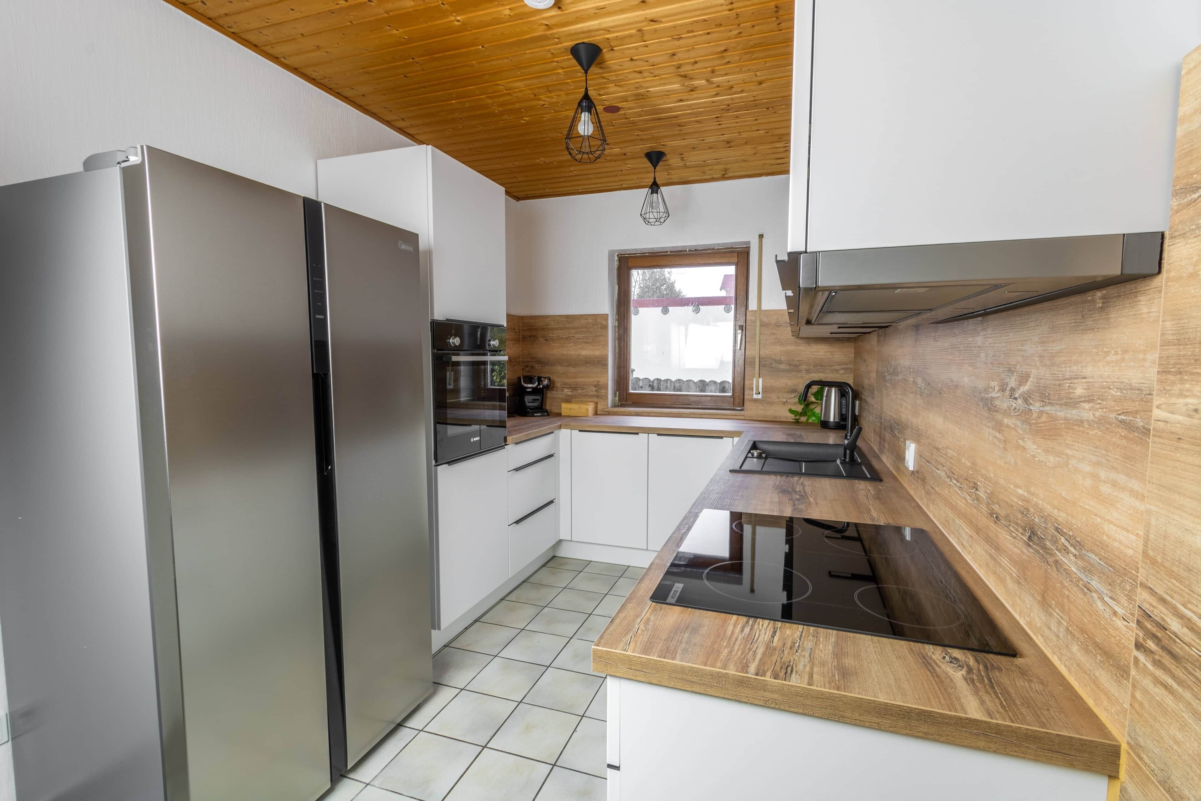 A modern kitchen with a stainless steel refrigerator, wooden cabinets, a built-in stove, and a window with a view.