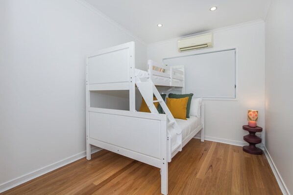 Una habitación pequeña y blanca con una cama litera, suelo de madera, un equipo de aire acondicionado y una pequeña mesa con una lámpara.