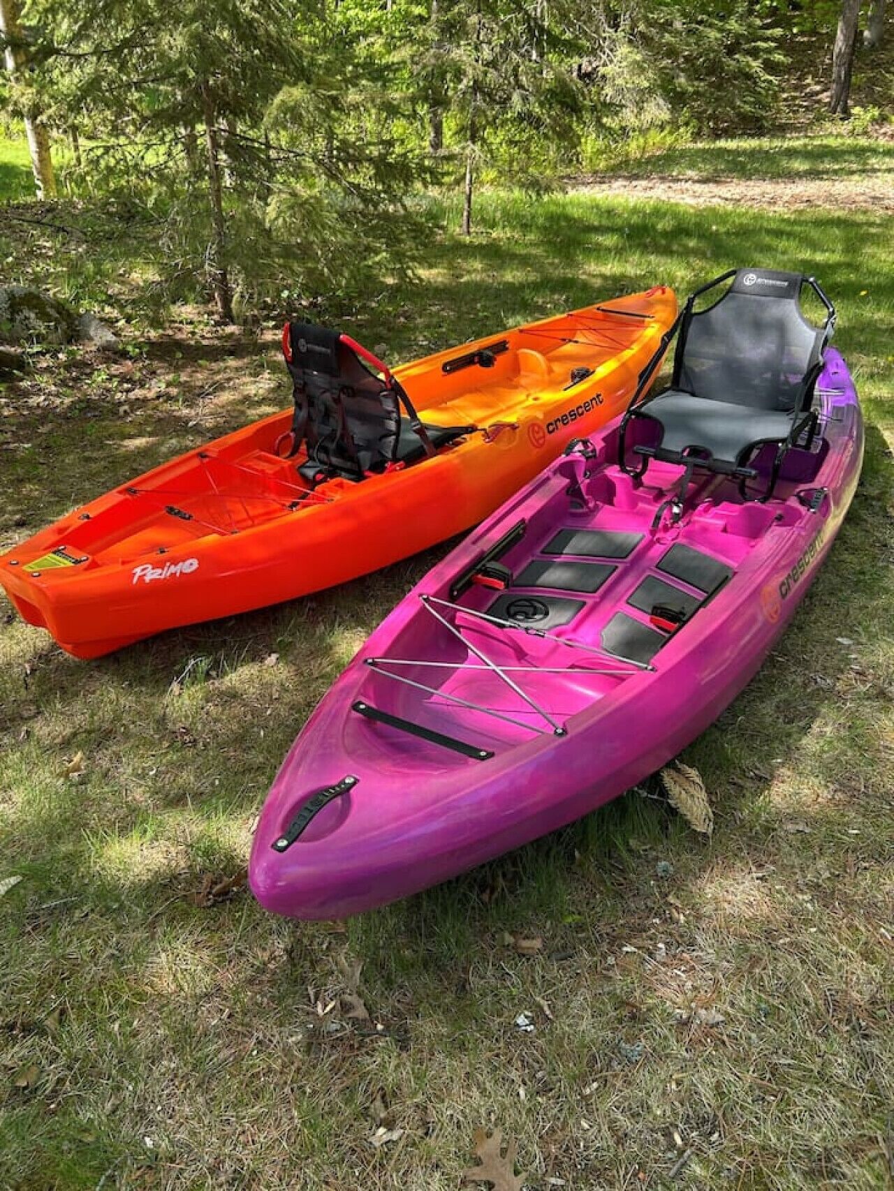 Deux kayaks, l’un orange et l’autre violet, sont garés sur l’herbe, avec des arbres en arrière-plan.