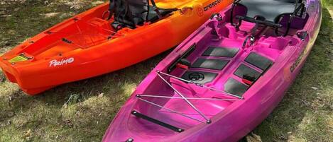 Deux kayaks, l’un orange et l’autre violet, sont garés sur l’herbe, avec des arbres en arrière-plan.