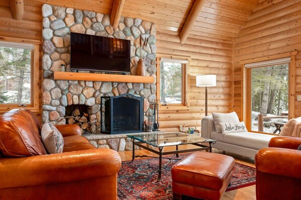 Interior acogedor de una cabaña de troncos con chimenea de piedra, mobiliario de cuero y grandes ventanales que ofrecen vistas al bosque.