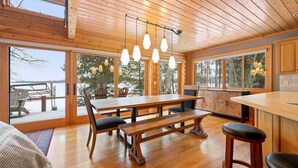 Interior de una cabaña de madera con comedor, cocina y ventanas amplias que ofrecen vistas a un paisaje nevado.