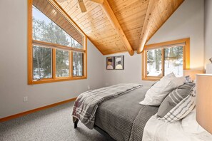Un dormitorio con techo inclinado, ventanas amplias, una cama con manta a cuadros gris y blanco, y vista a un bosque nevado.