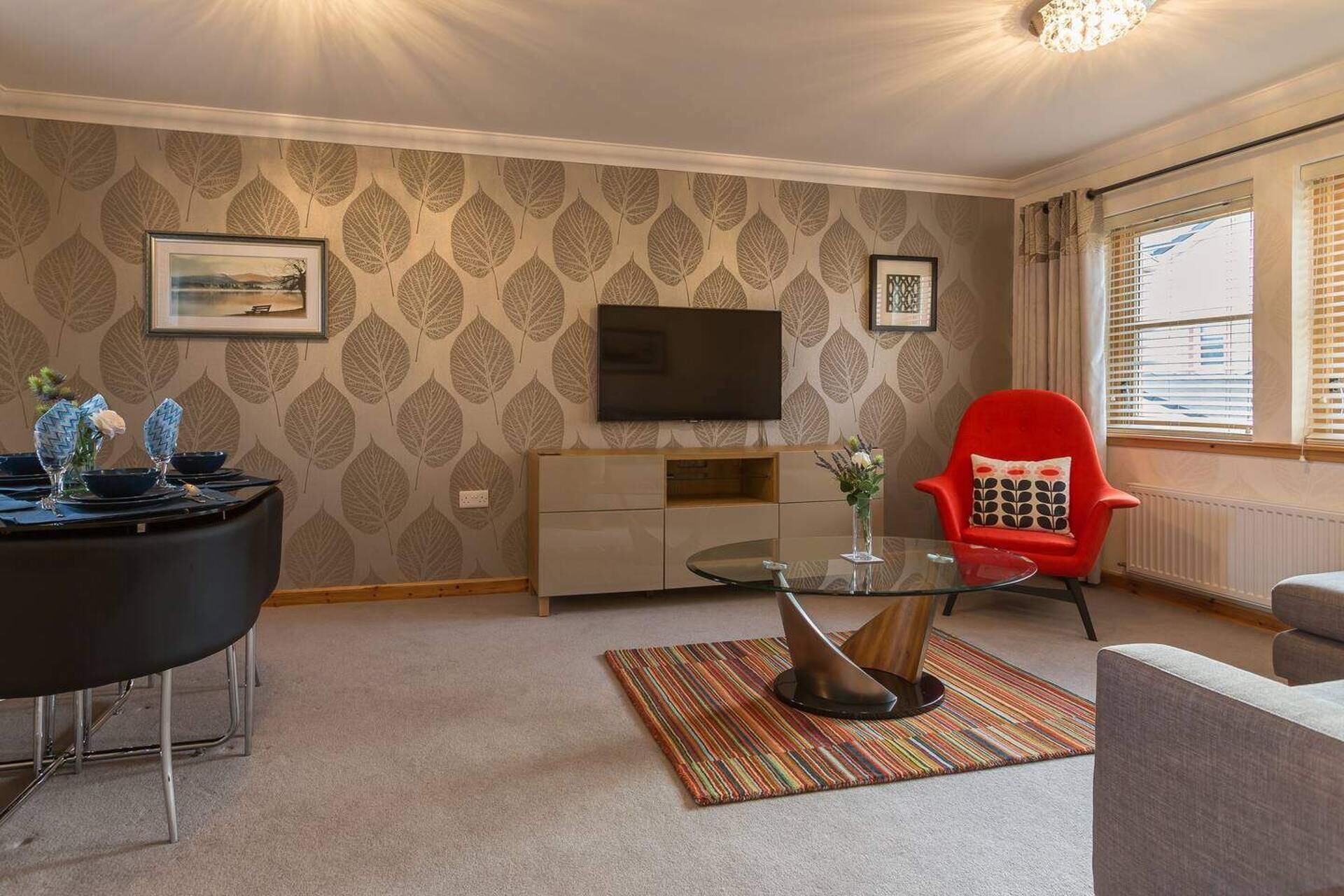 A living room with a flat-screen TV, a red chair, a glass coffee table, and a patterned carpet.