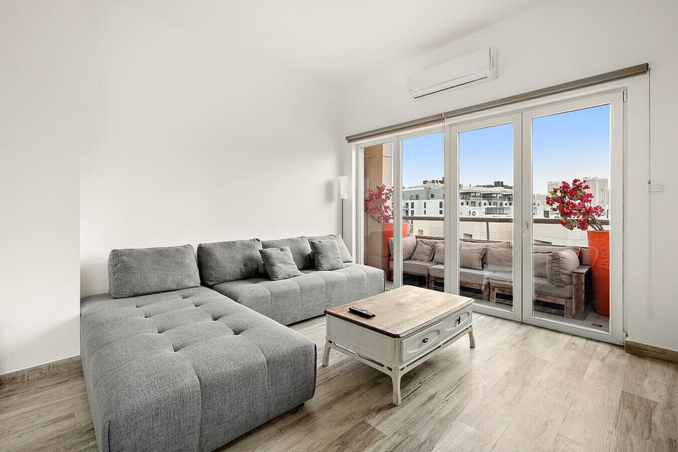 Un salon moderne avec un canapé d’angle gris, une petite table basse en bois et une porte-fenêtre coulissante donnant sur un balcon extérieur avec vue sur la ville.