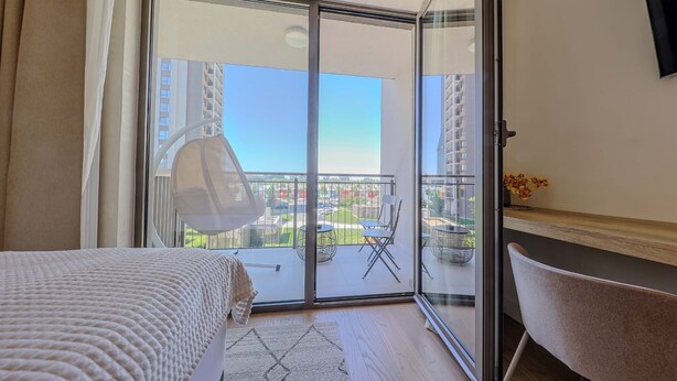 Habitación de hotel con cama, escritorio, una silla y balcón con vistas a edificios y vegetación.