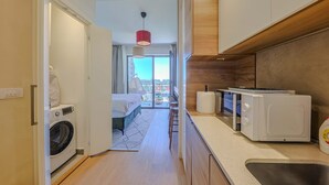 Habitación de hotel compacta con lavadora, cama, una pequeña kitchenette con microondas y vistas a la ciudad a través de un amplio ventanal.