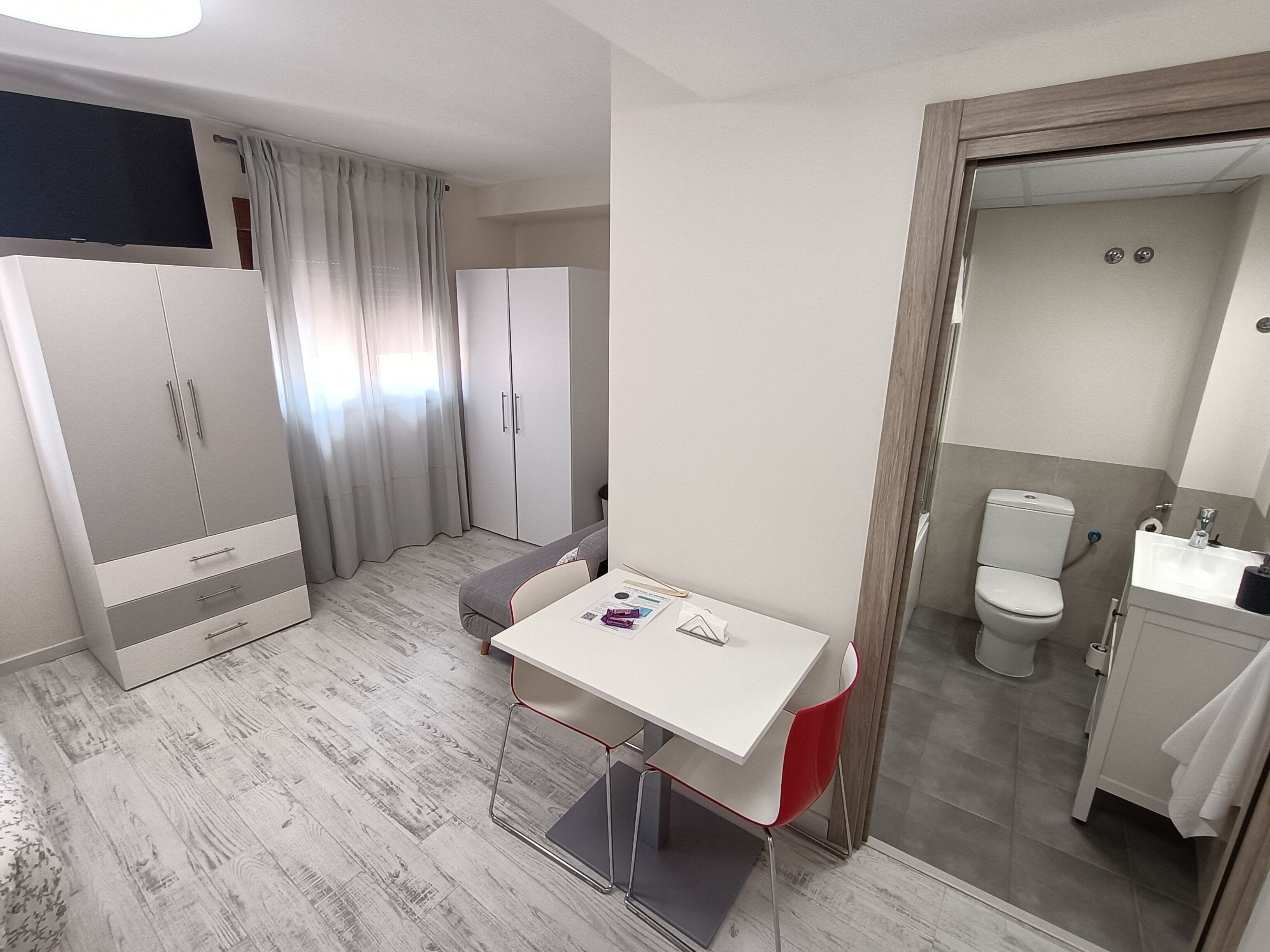 Une chambre d’hôtel compacte comprenant un lit, une petite table avec une chaise, une armoire, une salle de bain avec des toilettes et un lavabo, ainsi qu’un téléviseur fixé au mur.