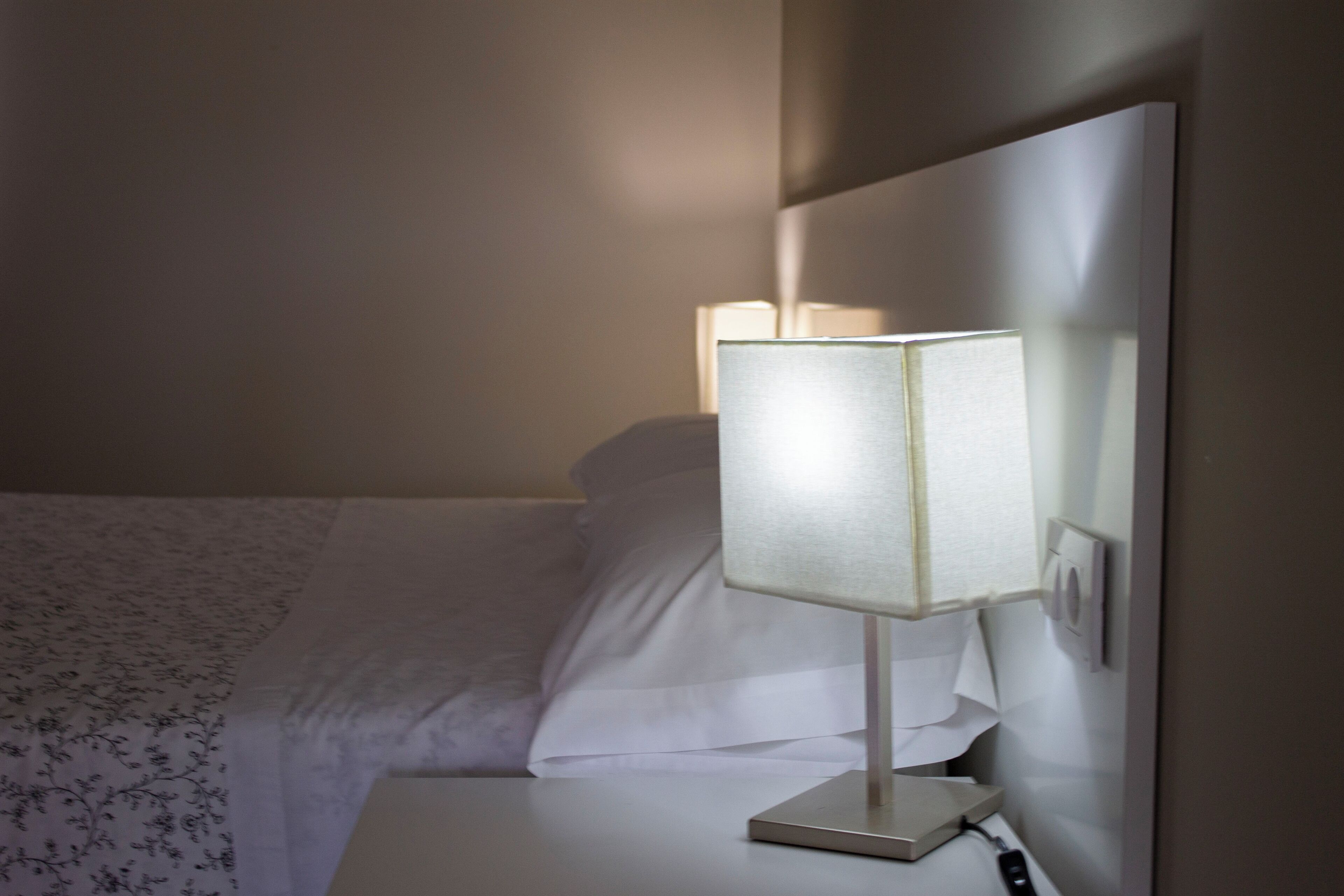 A neatly made bed with a floral-patterned blanket, a bedside table with a lamp, and a wall-mounted thermostat.