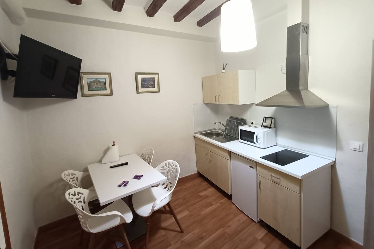A compact kitchen with a microwave, sink, and wooden cabinets. There is a dining table with chairs and a hanging light fixture.