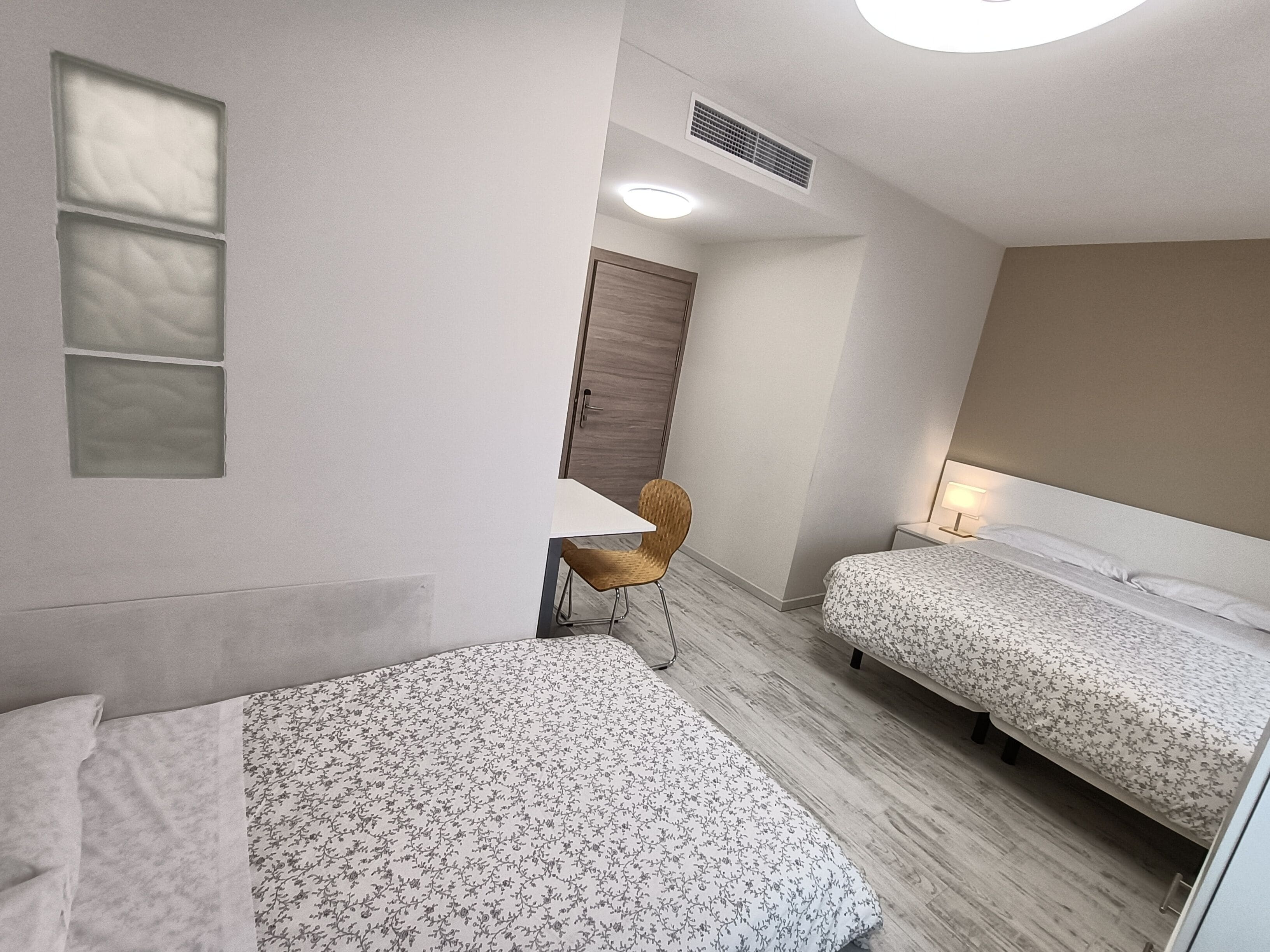 Une chambre d’hôtel avec deux lits, une chaise, une porte en bois et une table de chevet avec une lampe.