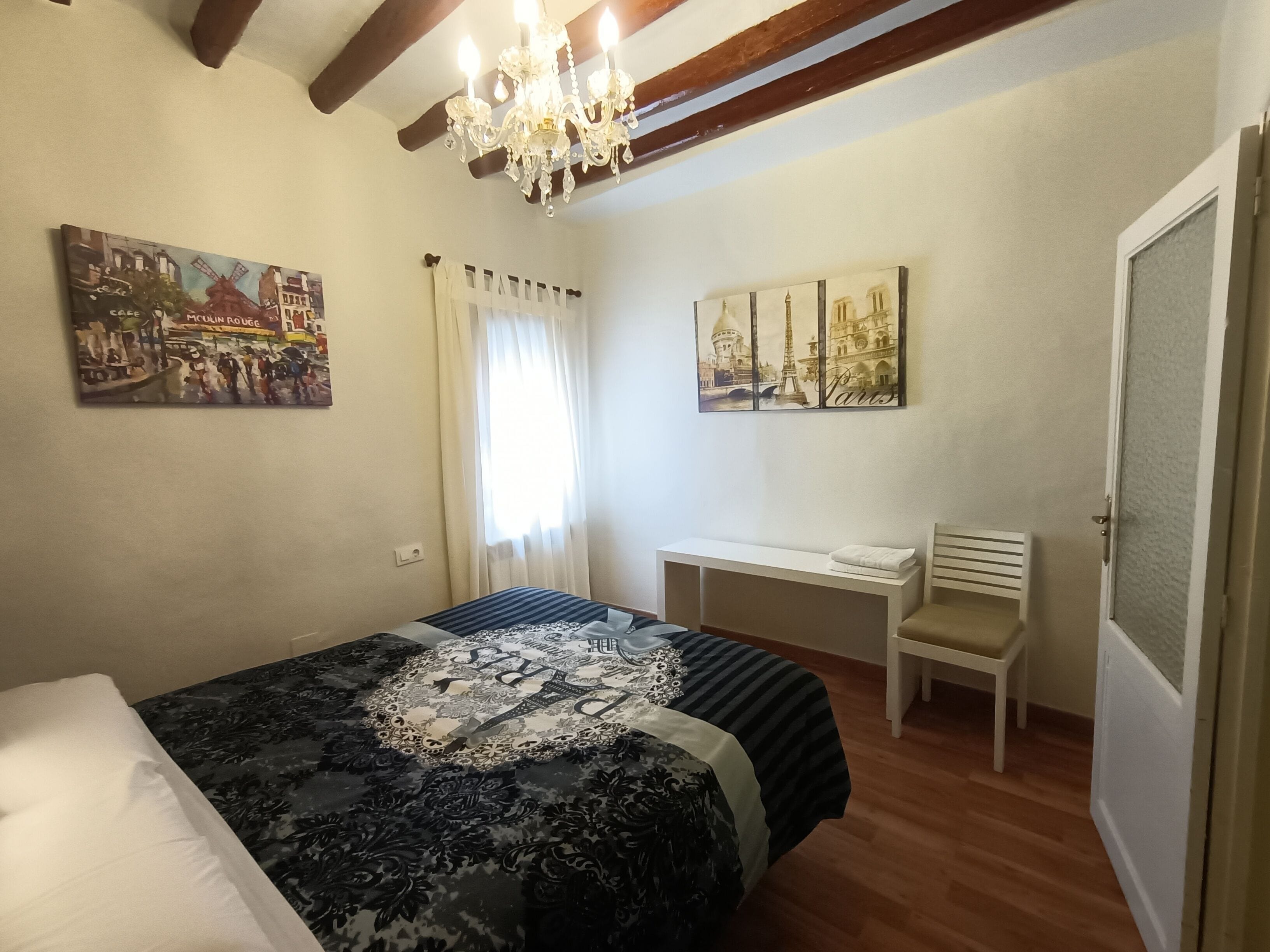 Un dormitorio con cama, escritorio y silla. Hay cuadros en las paredes y una araña.