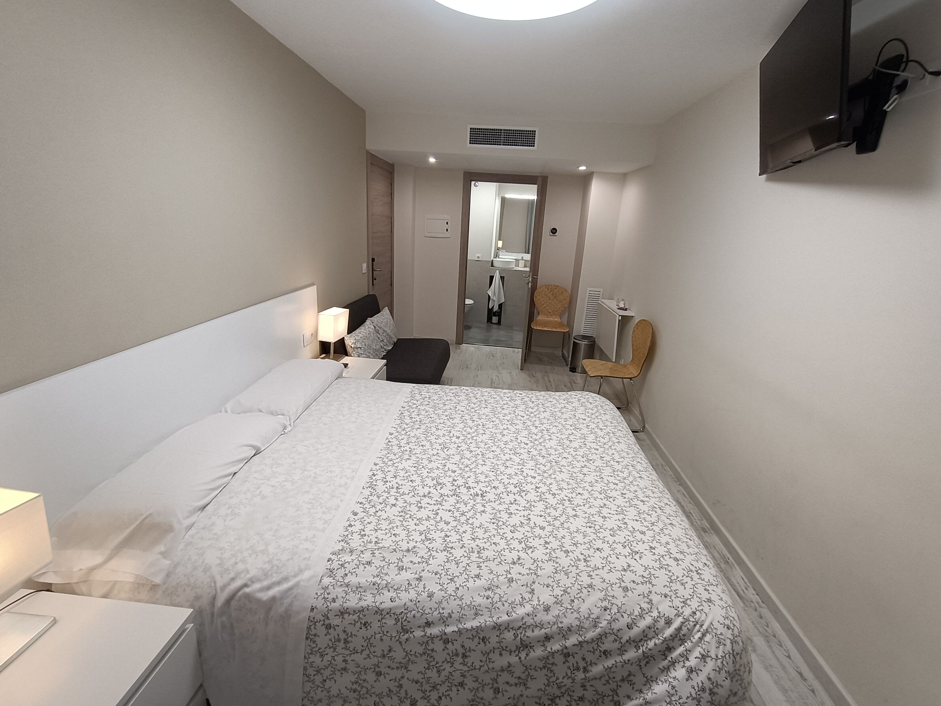 A hotel room with a bed, bedside table, lamp, chair, and a TV mounted on the wall.