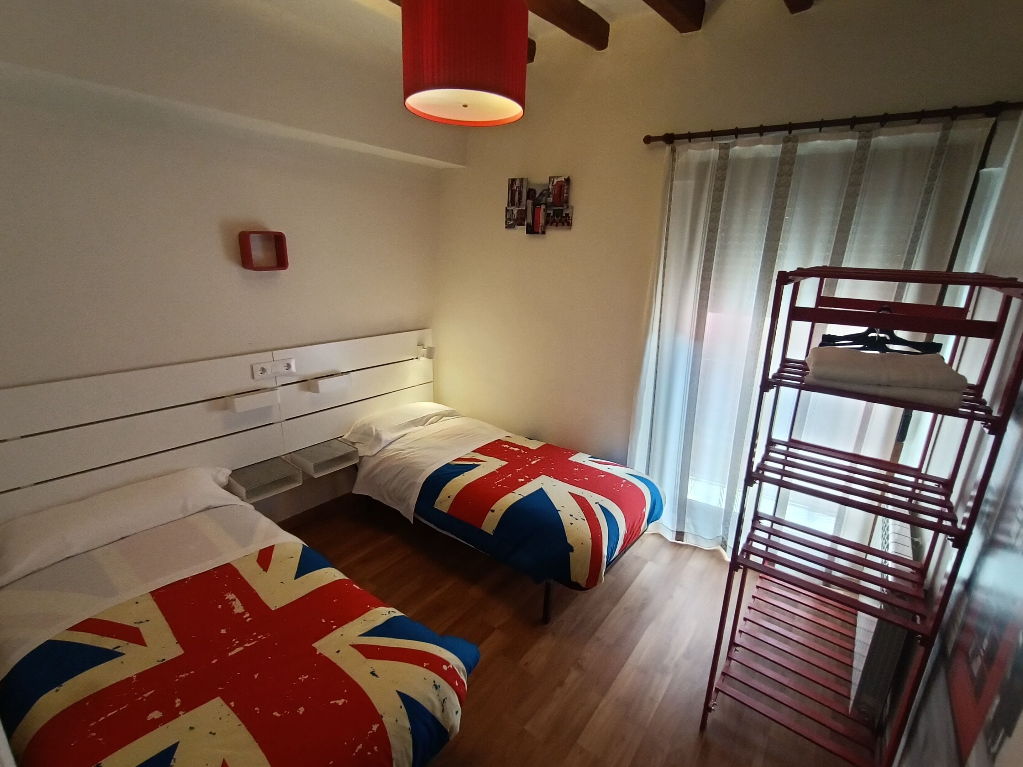 A room with two beds, a bunk bed, wooden flooring, a red hanging light, a window with curtains, and a wall-mounted shelf.
