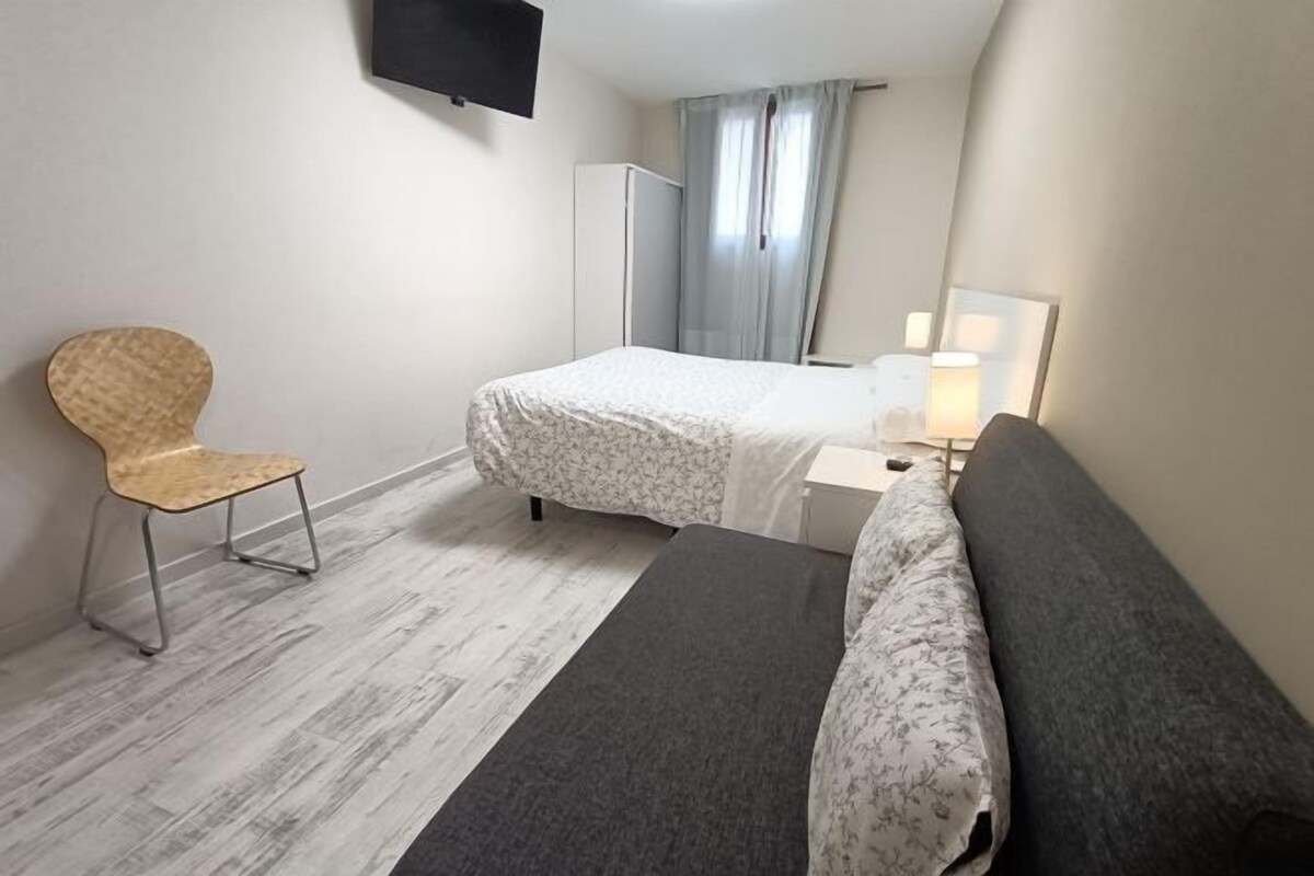 A small, neatly arranged room with a bed, a chair, a desk with a lamp, and a wardrobe.