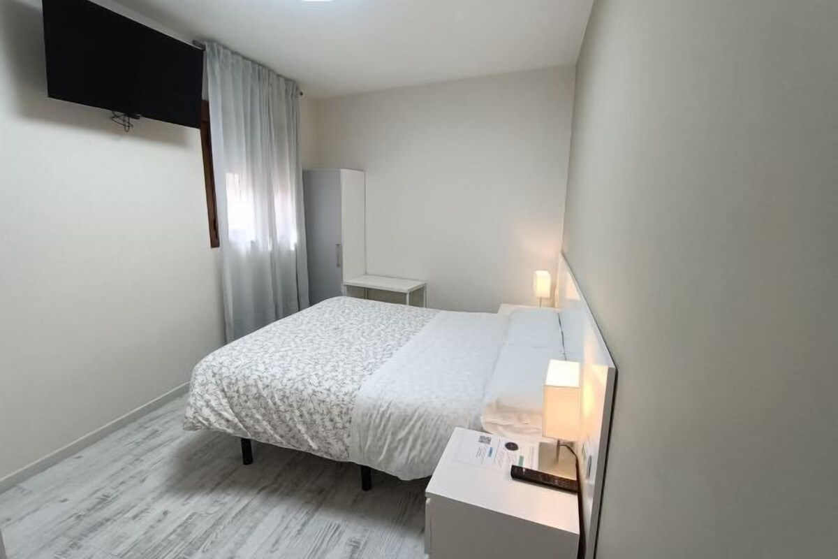A bedroom with a bed, bedside table, lamp, and a flat-screen TV mounted on the wall.