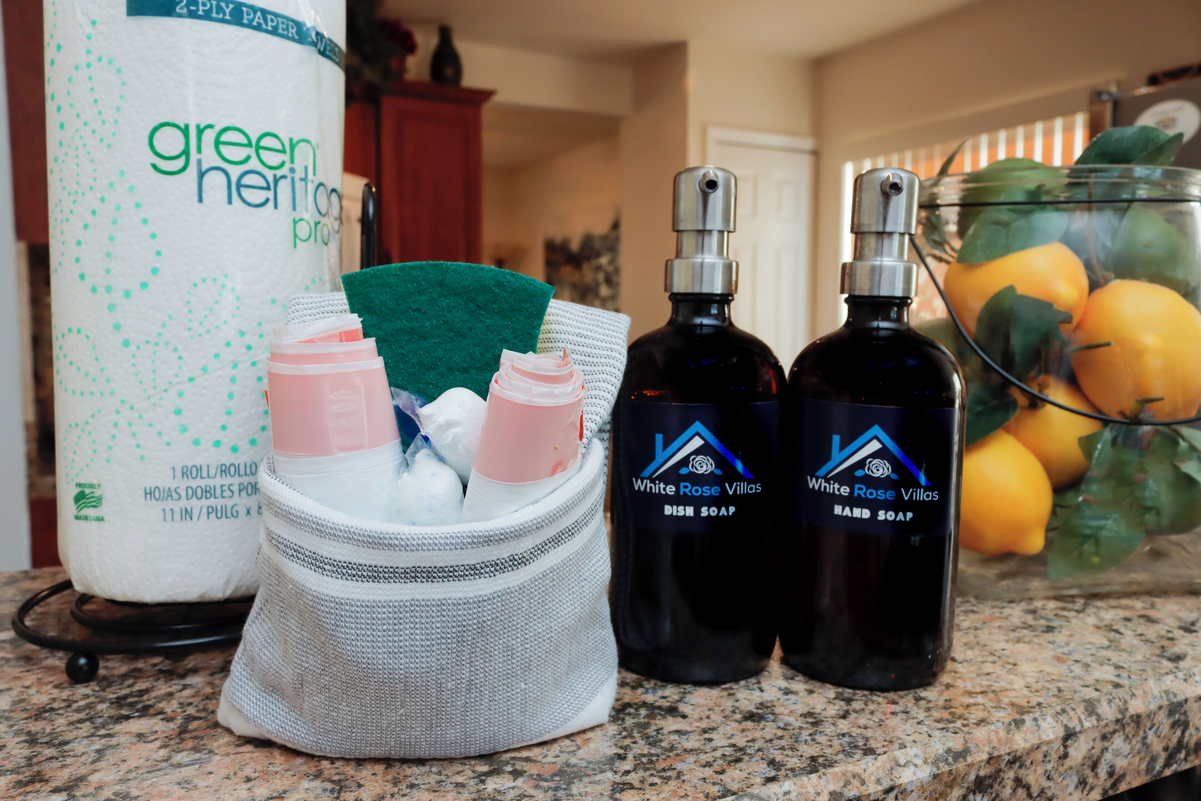 A kitchen counter with a 2-ply paper towel roll, a mesh container with soap and sponge, and two bottles of dish soap.