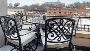 Un balcon avec une table ronde et quatre chaises, donnant sur un paysage urbain avec des bâtiments et un ciel dégagé.