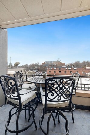 A balcony with a round table and four chairs, overlooking a cityscape with buildings and a clear sky.