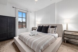 A bedroom with a large bed, a dark wardrobe, a window with curtains, and a bedside table with a lamp.