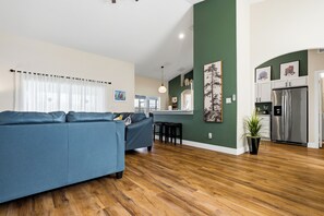 Una sala de estar moderna con un sofá azul, suelo de madera y una cocina con refrigerador de acero inoxidable.