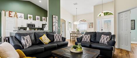 A living room with two black leather sofas, a coffee table, and a kitchen in the background.