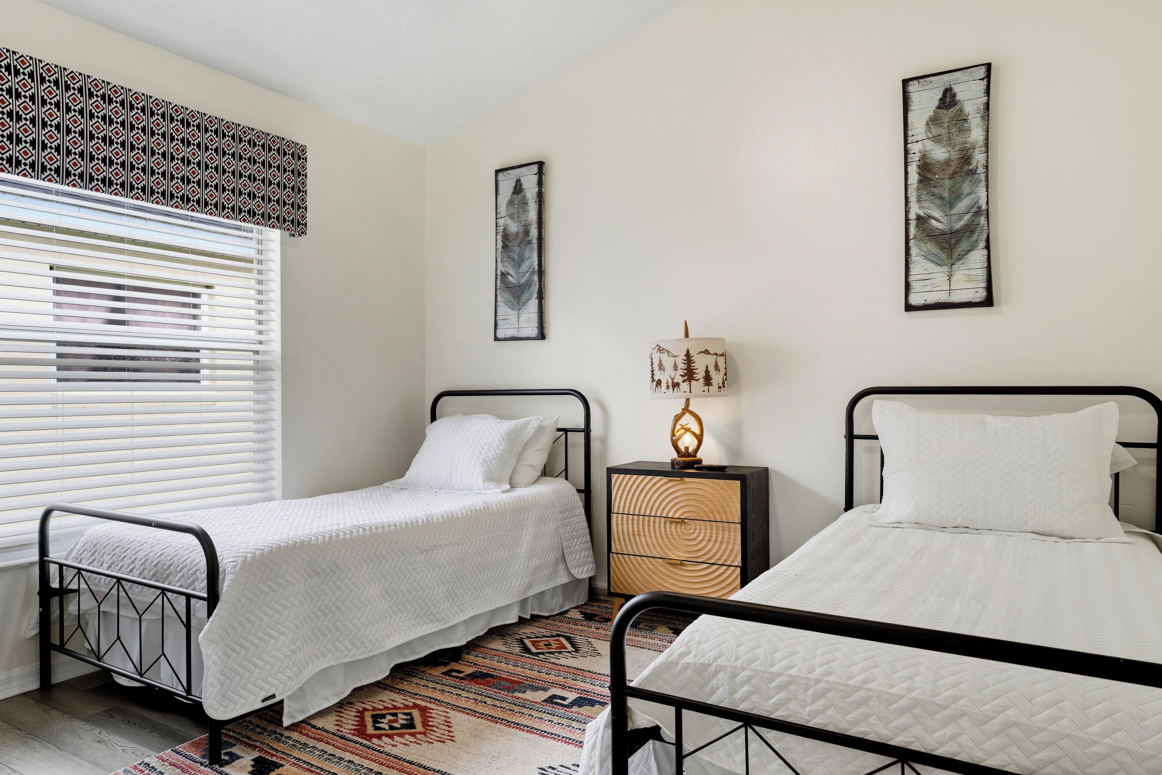 A bedroom with two single beds, a bedside table with a lamp, a patterned rug, and a window with blinds.
