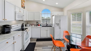 A modern kitchen with white cabinetry, a built-in oven, a microwave, a refrigerator, and a dining area with orange chairs. - Disney Area, Pool, Game Room, Perfect for Families (Kissimmee)