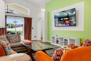 A modern living room with a flat-screen TV mounted on a green wall, a glass coffee table, and a sofa with colorful cushions. - Perfect Family Disney Escape: Pool & Games (Kissimmee)