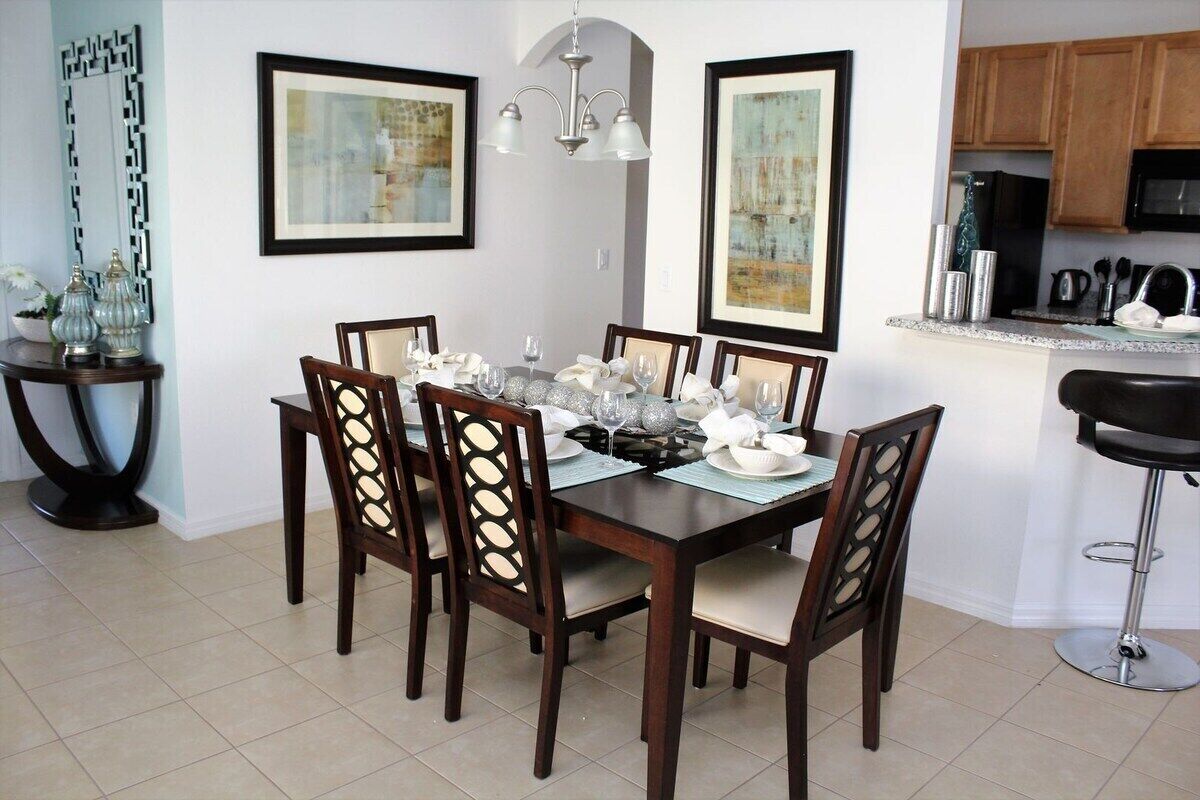 Une salle à manger avec une table en bois foncé, dressée pour six personnes, entourée de chaises assorties. Les murs sont ornés d’œuvres d’art encadrées, un lustre est suspendu au-dessus de la table, et on aperçoit un coin cuisine sur la droite.