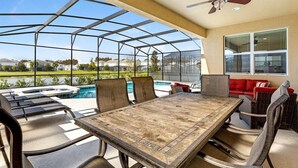 A covered outdoor area with a table and chairs, a ceiling fan, and a view of a swimming pool and landscape. - Magical Disney Villa • Sunny Pool, Spa & Game Room (Kissimmee)