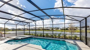 A swimming pool with a glass enclosure, surrounded by a concrete deck and a view of a lake and houses in the background. - Magical Disney Villa • Sunny Pool, Spa & Game Room (Kissimmee)