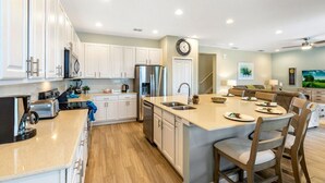 A modern kitchen with white cabinets, a stainless steel refrigerator, a dishwasher, and a large island with seating. - Magical Disney Villa • Sunny Pool, Spa & Game Room (Kissimmee)