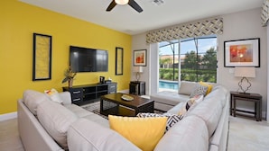A living room with a yellow wall, a ceiling fan, a flat-screen TV, a sofa, a coffee table, and a window with a view of greenery. - Luxury Resort Villa with Pool, Spa – Perfect for Families! (Davenport)