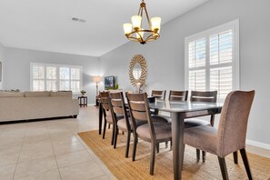 A dining area with a glass table, six chairs, a chandelier, and a large window with blinds. - Spacious 5BR West Haven Villa Near Disney | Private Pool, Spa & Game Room (Davenport)