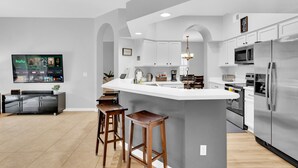 Une cuisine moderne dotée d’un grand îlot central, de meubles blancs, d’appareils électroménagers en acier inoxydable et d’un téléviseur à écran plat fixé au mur.