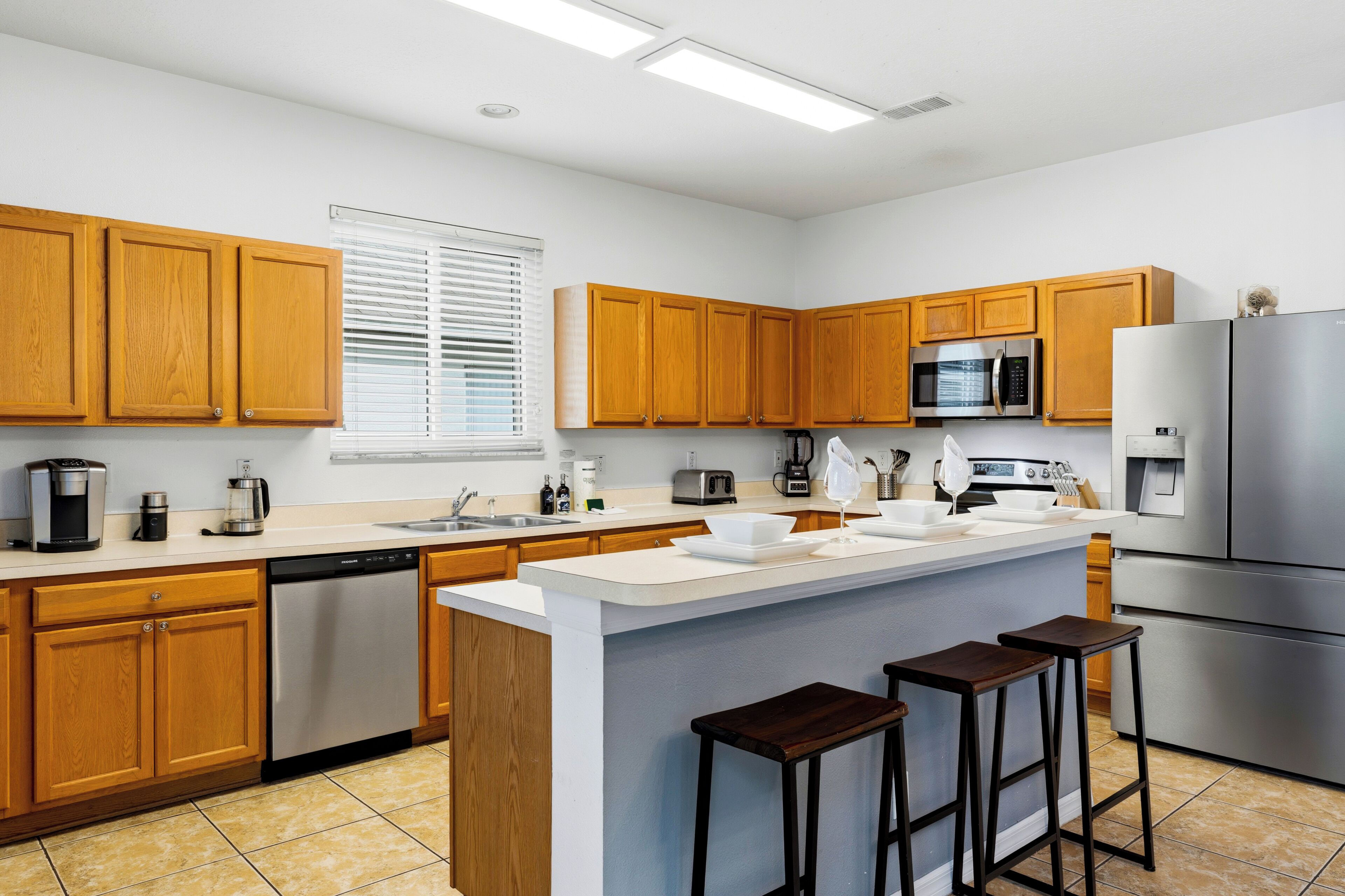 A modern kitchen with wooden cabinets, stainless steel appliances, a central island with stools, and a window with blinds.
