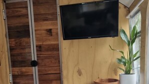 A compact living space with a wooden door, a flat-screen TV mounted on the wall, a potted plant, and a small table with a bowl and placemats. - Escape to a unique retreat with Miss Beddy, recently renovated Bedford bus  (Mount Helen)