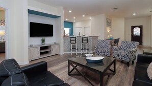 A modern living room with a flat-screen TV, black leather sofas, a glass coffee table, patterned armchairs, and a ceiling fan. - Stunning 4BR Disney Villa w/ Pool & Arcade (Clermont)