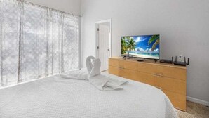 Un dormitorio con cama, televisor sobre una cómoda de madera y ventana con cortinas traslúcidas.