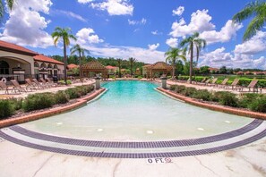 A large outdoor swimming pool surrounded by lounge chairs and palm trees, with a clear sky above.