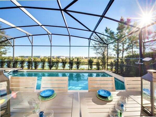 An outdoor swimming pool with a glass enclosure, surrounded by wooden deck chairs and tables, overlooking a scenic landscape.