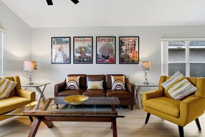 A modern living room with a brown leather sofa, yellow armchairs, a glass coffee table, and framed posters of theme parks.