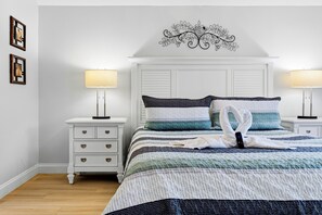 A neatly made bed with a striped blanket, two pillows, and a swan-shaped pillow. There are two bedside tables with lamps, a wall-mounted decorative piece, and a white dresser with drawers.